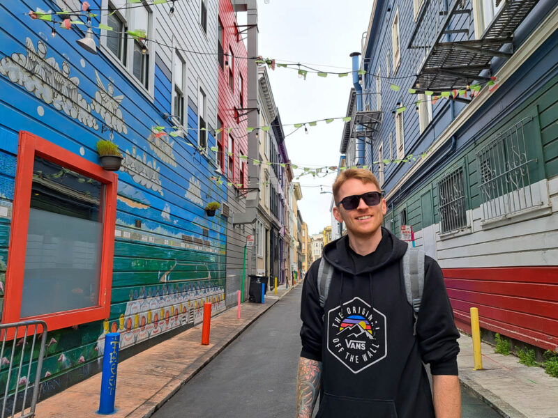 A man wearing a black hoodie and sunglasses standing in the middle of a quiet laneway in Little Italy, San Francisco with colorful buildings on either side in the colours of the Italian flag: red, white and green.