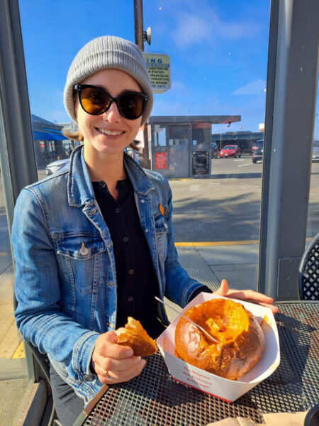 A woman with a giant smile on her face wearing a beanie, sunglasses and a denim jacket sitting outside eating a sourdough bread bowl soup.