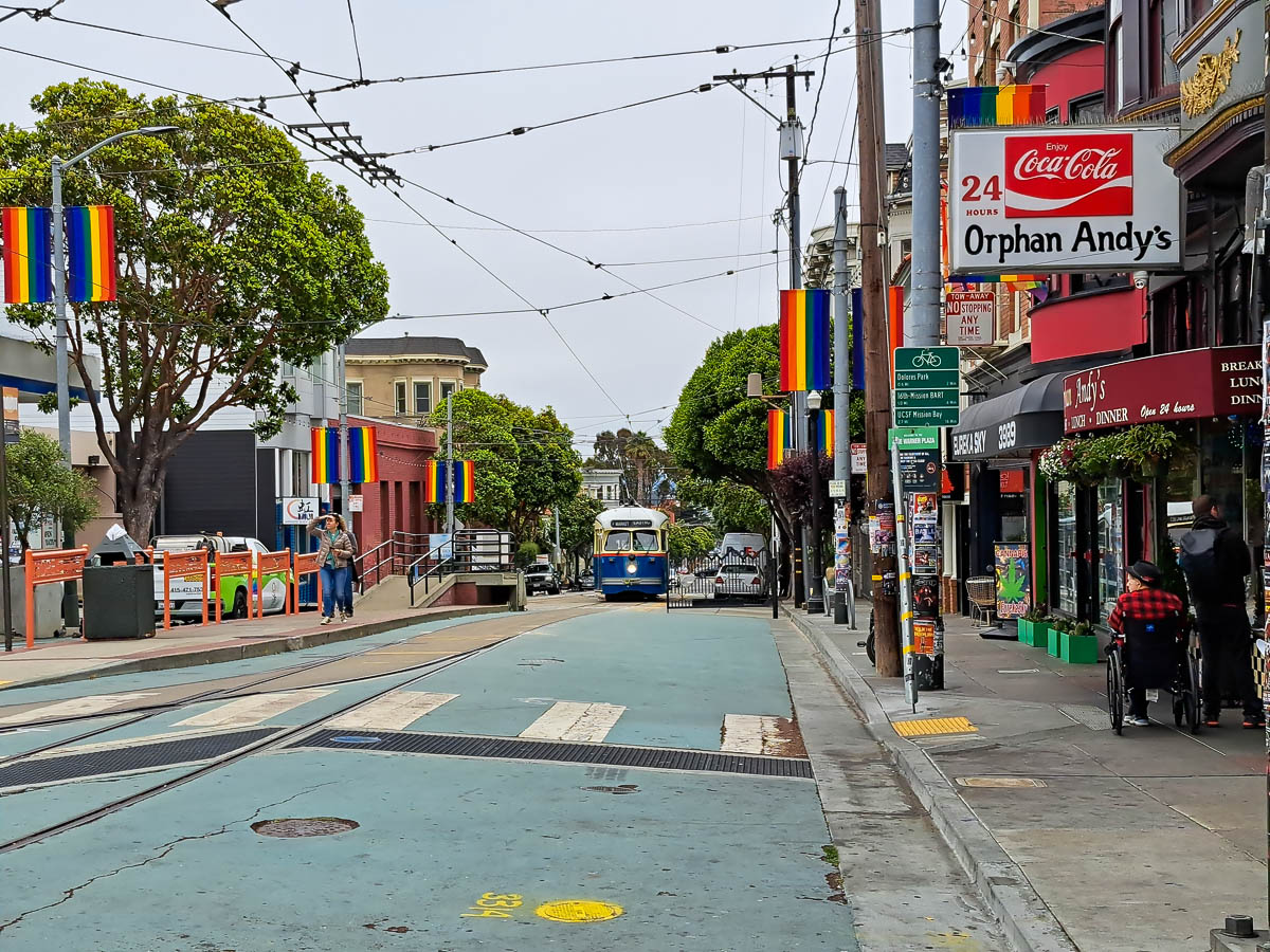 Castro District in San Francisco with a tram approaching, rainbow flags hanging off of buildings, and people milling about.