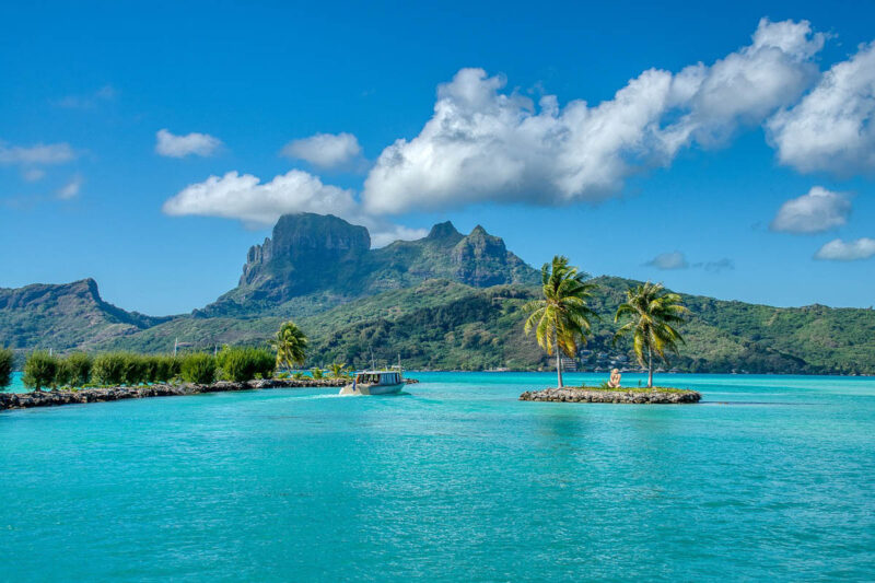 A dream romantic destinations vacation location of the island of Bora Bora with turquoise water, bright blue skies, fluffy white clouds, and lush tree and flora lined mountain