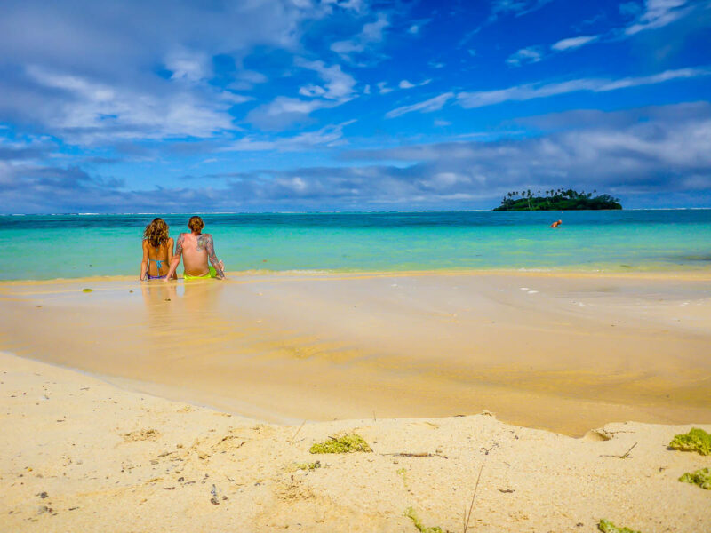 A man and woman sitting in shallow turquoise water on light sand beach enjoying a romantic destinations getaway in the Cook Island