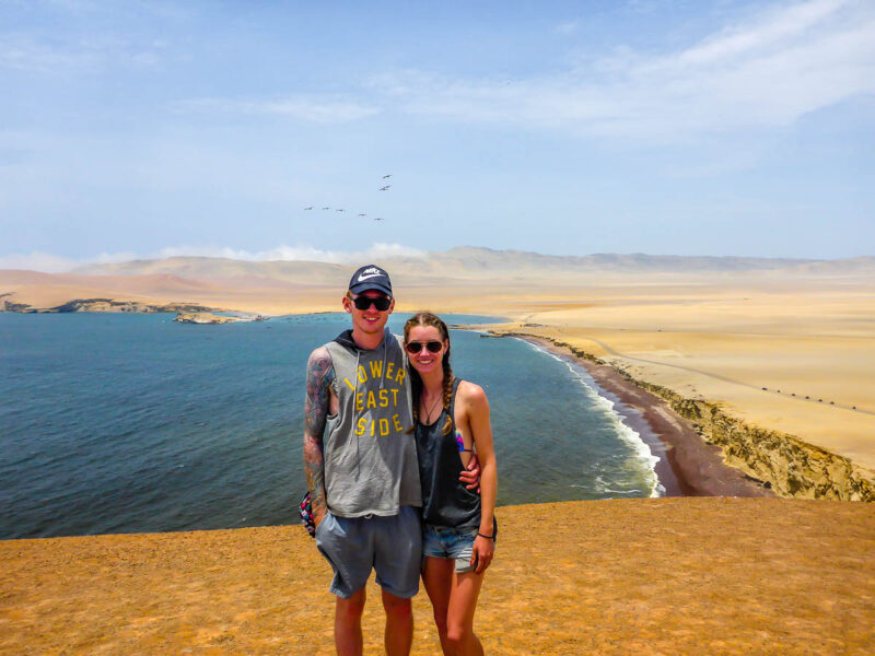 A man and woman standing on a rugged cliff overlooking rich natural landscapes with multi-colored sand, deep blue water and blue sky