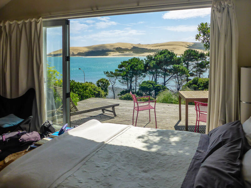 View from the side of a bed looking out double doors leading to a deck overlooking a bright blye lake