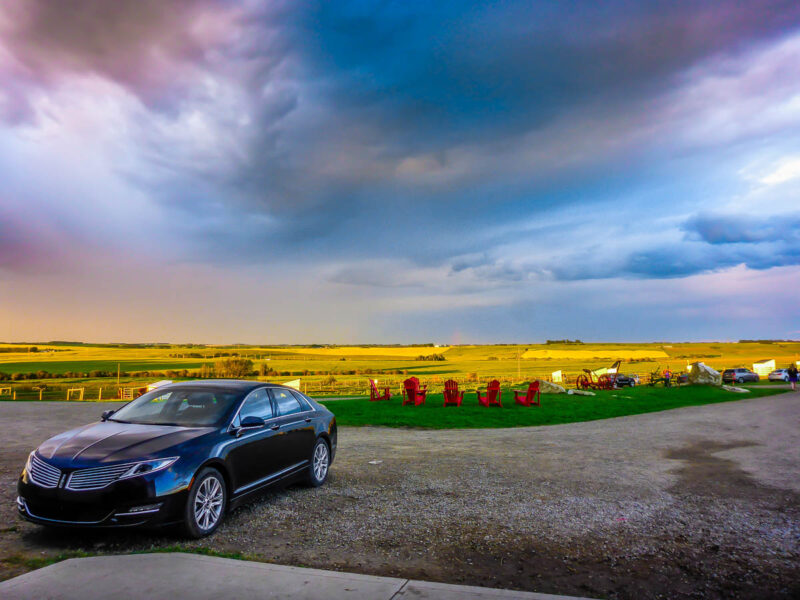 View of a car parked near a field with 5 red deck chairs, sunshine and moody colorful clouds