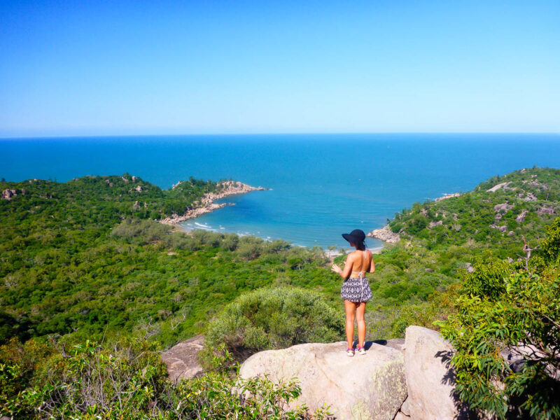 A woman standing on top of a boulder overlooking greenery and an ocean bay