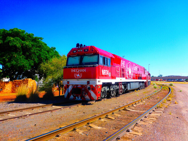 A big red train, the Ghan, traveling on sand and dirt in the Australian Outback with a tree and blue skies in the background