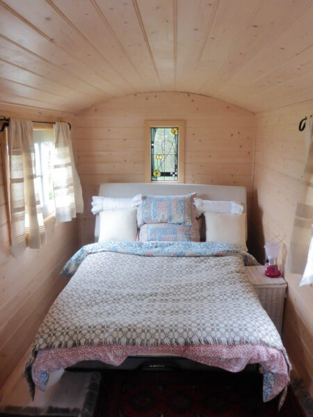 A double bed with light colored linens in a small caravan with light beige wood panels for the walls and ceiling, a great budget travel tip to stay in unique places