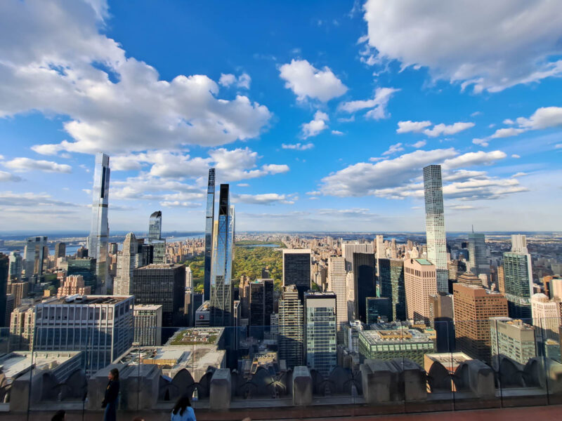 A view of New York City on a bright, clear, sunny day with blue skies, fluffy clouds and tall skyscraper buildings in the background. 