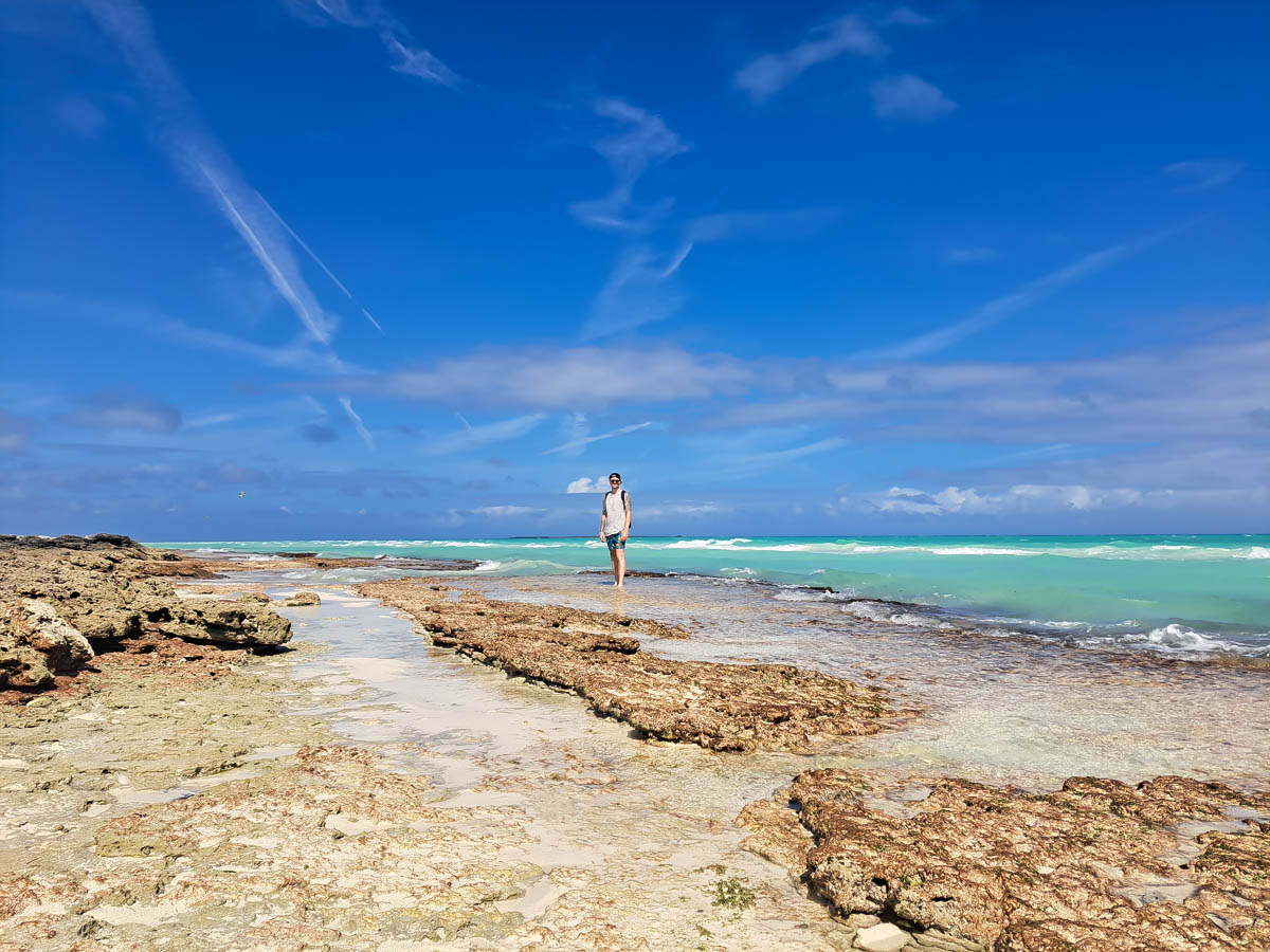 A man standing in on a sandy and rocky beach with lava rocks, clear turquoise ocean water, wispy clouds and deep blue skies.