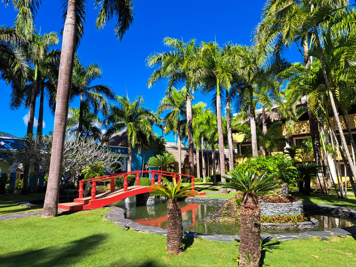 A red bridge over a small pond at an all-inclusive resort in the Dominican Republic with green grass, palm trees, the resort and blue sky