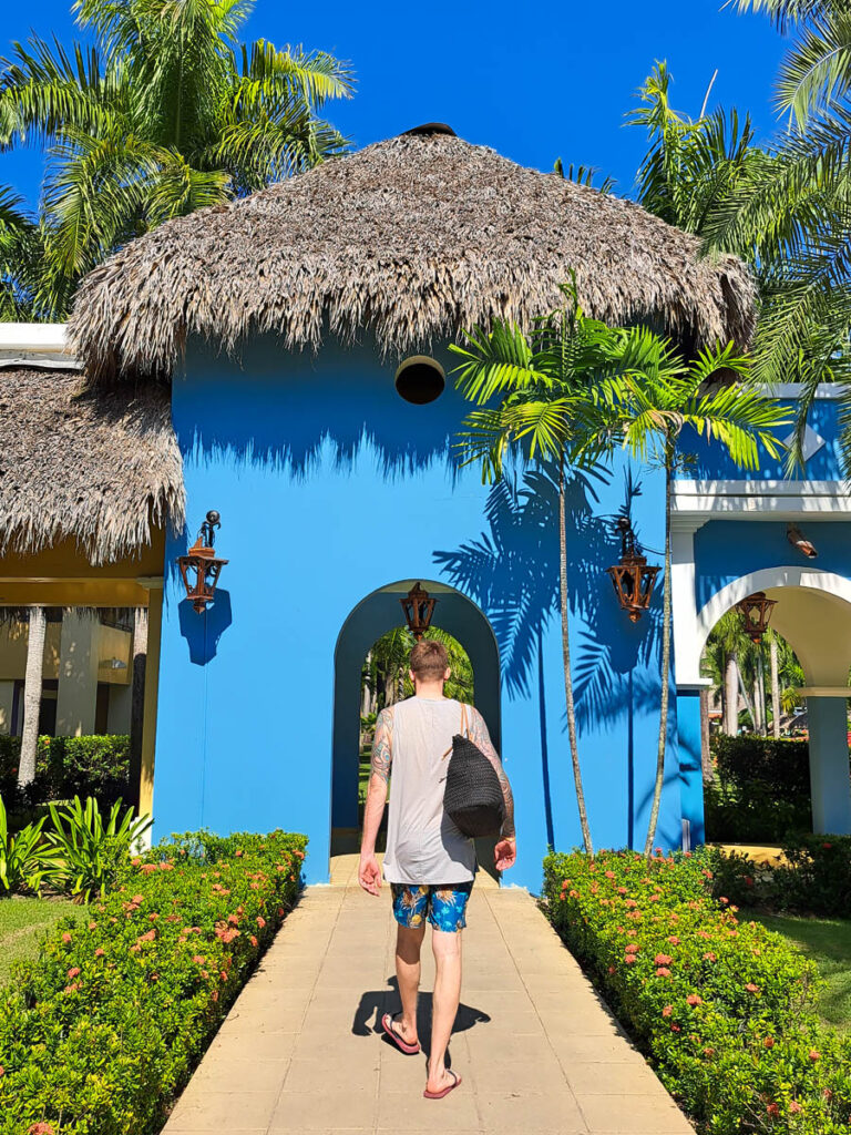 A man walking away from the camera carrying a beach tote walking towards a bright blue archway at a tropical resort