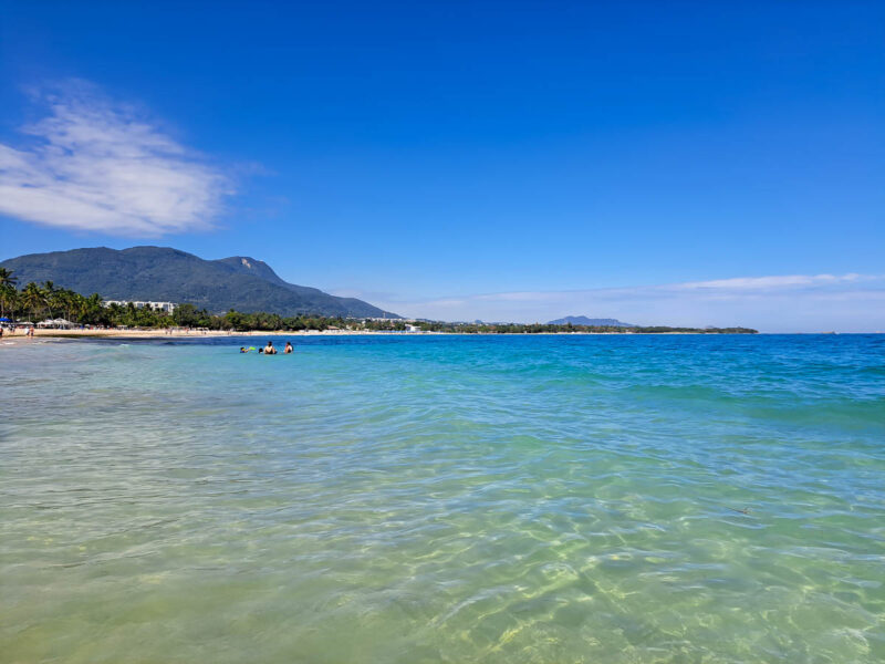 Clear, turquoise waters, blue sky with a couple fluffy clouds, a sandy beach and mountains in the background