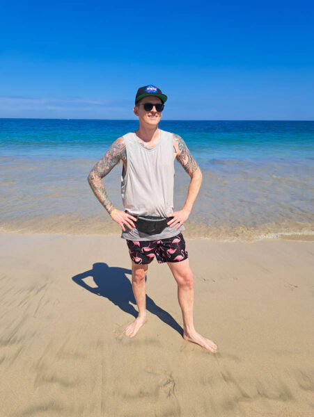 A man posing on a sandy beach in the Dominican in front of turquoise blue ocean waters wearing a sparkly black fanny pack