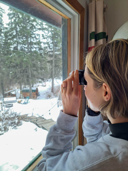 A woman looking out the window with binoculars, looking out to a winter forest with snow, sparse pine and birch trees, a wood-barrel sauna and wooden steps.