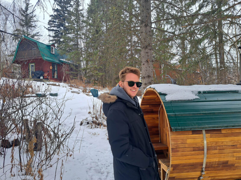 A man in a winter coat standing next to a wood barrel sauna in the winter with pine and birch trees all around and a red-brown barn in the background up a small hill.