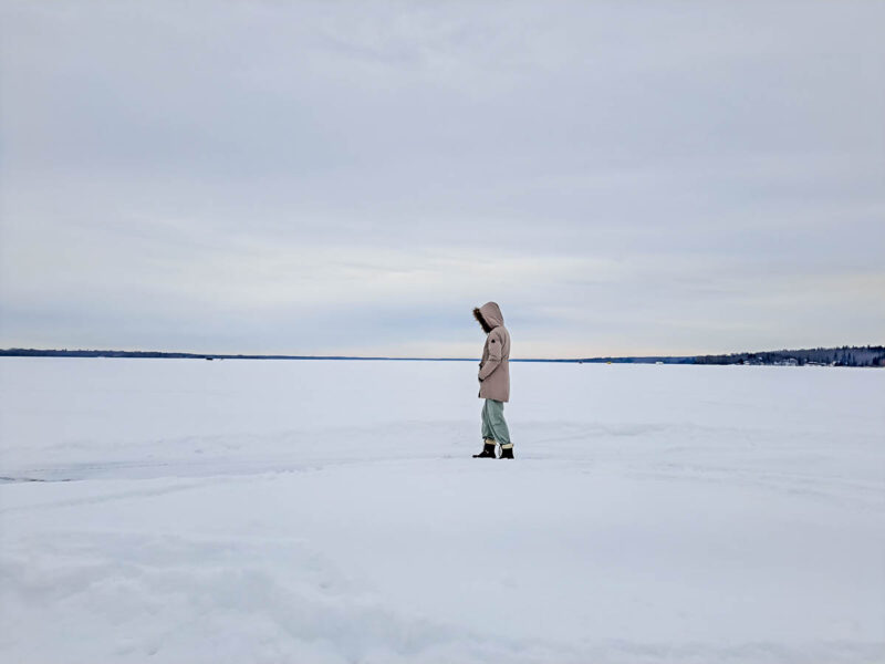 A woman walking on the frozen Lac la Nonne in Birch Cove, Alberta with white ice, snow, and white cloudy sky in the background.