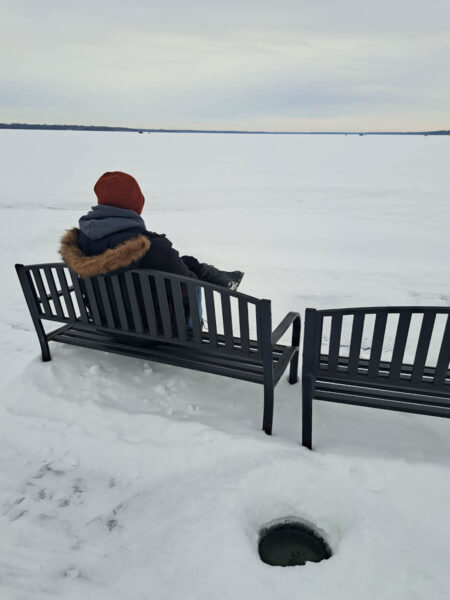 The backside of a man sitting on a bench overlooking a frozen lake with ice fishers off in the distance, white ice and snow, and why cloudy sky.