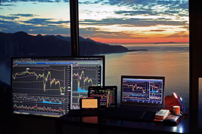 Multiple computer screens looking at financial stock market indexes and charts, with a view outside a window of a body of water and sunset