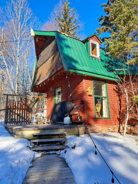 A red-brown barn with bright green roof, wooden steps leading up to it, fairy lights hanging off of it, set in a winter forest and bright blue skies