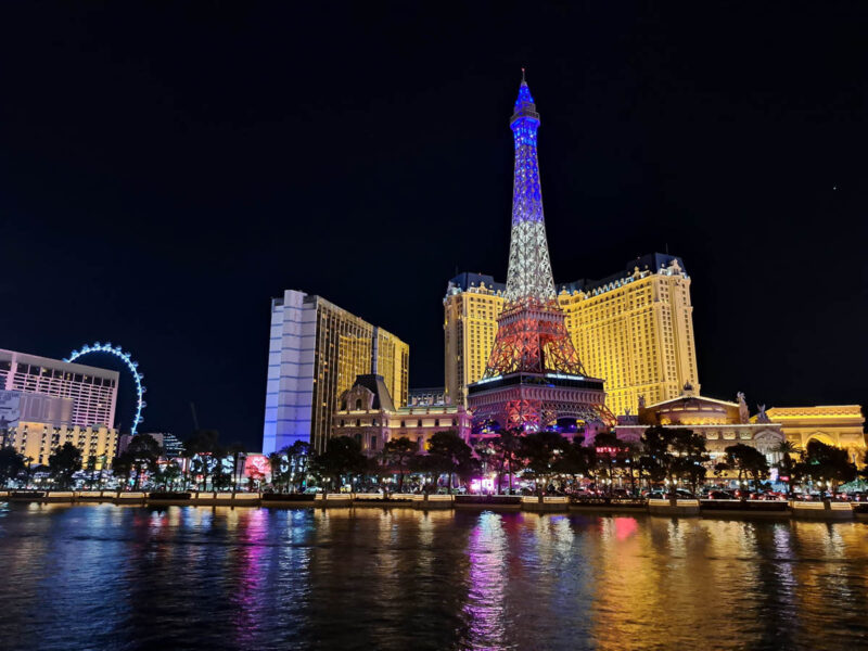 Las Vegas strip at nighttime with an imitation Eiffel Tower lit up the colors of the French flag, a big lit up ferris wheel, tall hotels and other buildings in the background with colorful lights bouncing off the reflection of water.