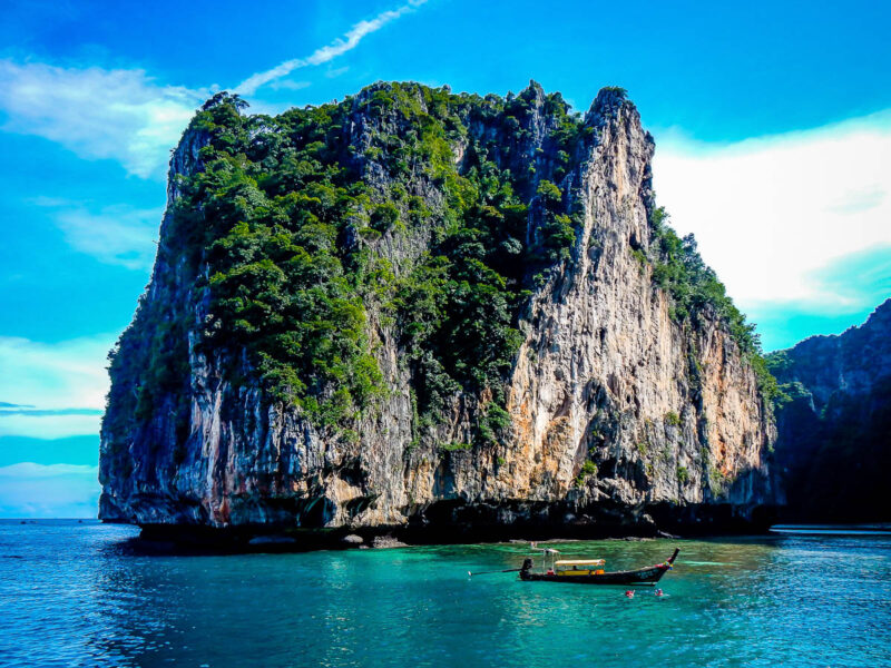 Rock island formation in Maya Bay, Thailand with a small boat in front, crystal clear waters and bright blue skies