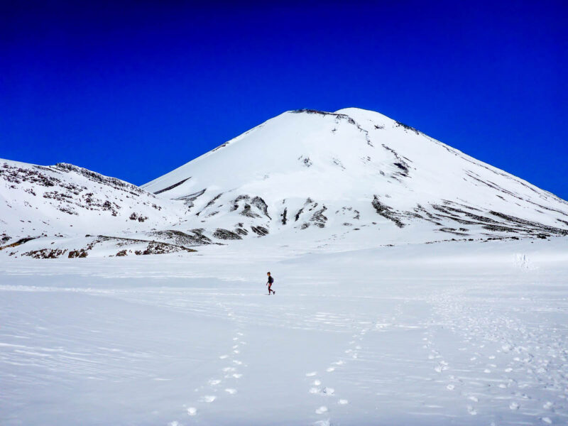 A woman in the distance walking in the snow with a snow covered mountain behind her and deep blue skies.