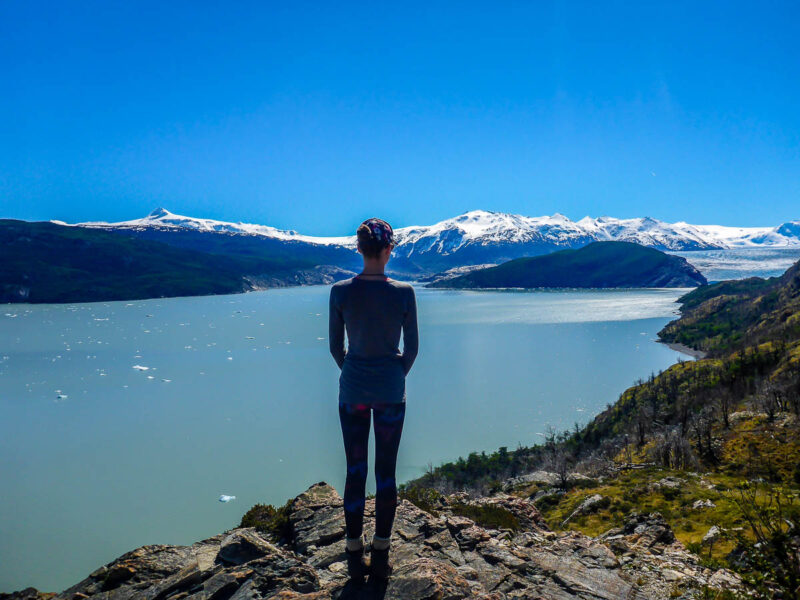 A woman with her back to the camera standing on the side of a mountain looking at a glacial lake with turquoise water, and glacial mountains in the background. Bright blue sky.