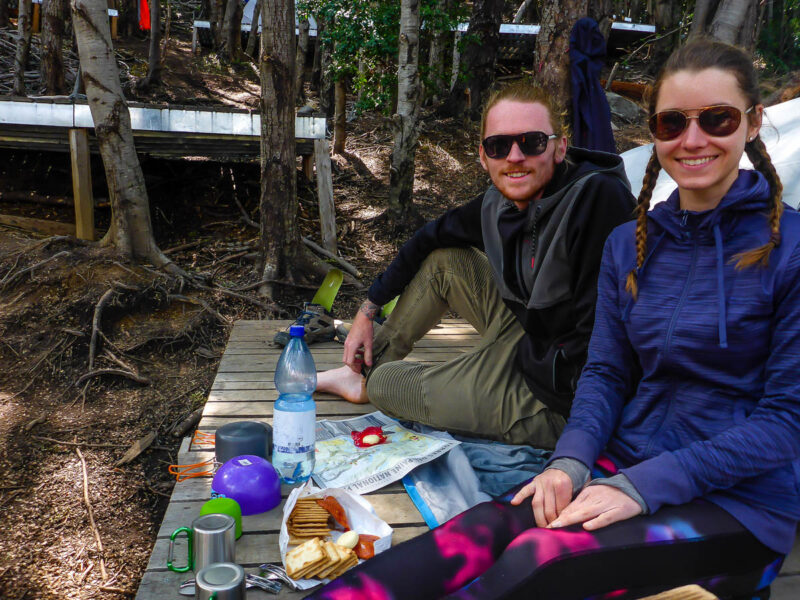 A couple sitting on a wooden platform in the middle of the forest eating crackers and cheese surrounded by other cooking equipment