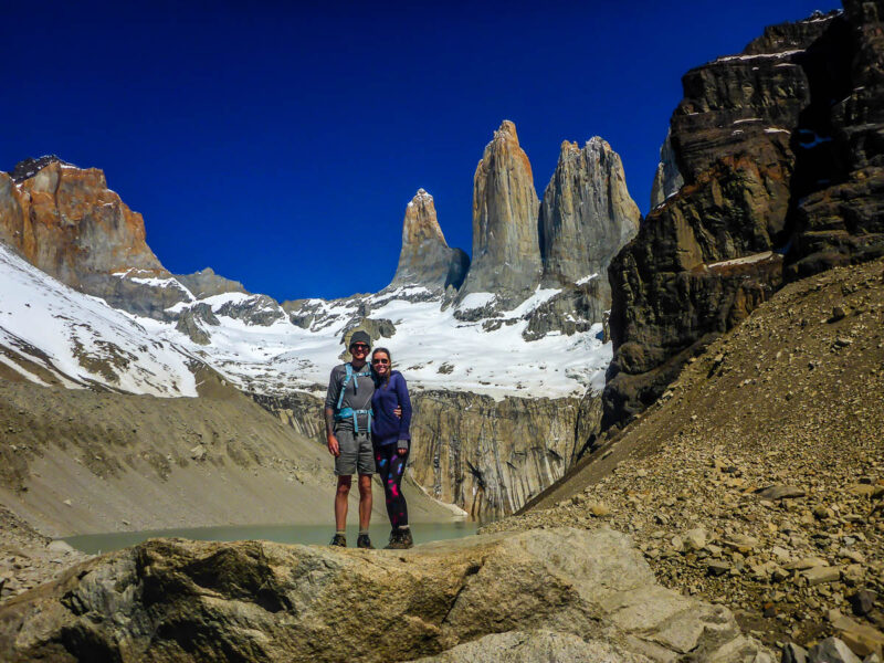 A couple standing on a large boulder in front of a small glacial lake and glacial mountains covered in snow behind them. Three tall mountains shoot out in the background known as Fitz Roy.