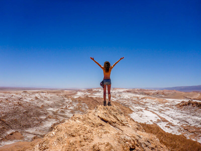 A woman standing with her back towards the camera on top of a hill overlooking the Atacama Desert in Chile. The sand is different shades of browns, reds, beiges and whites with deep blue sky.