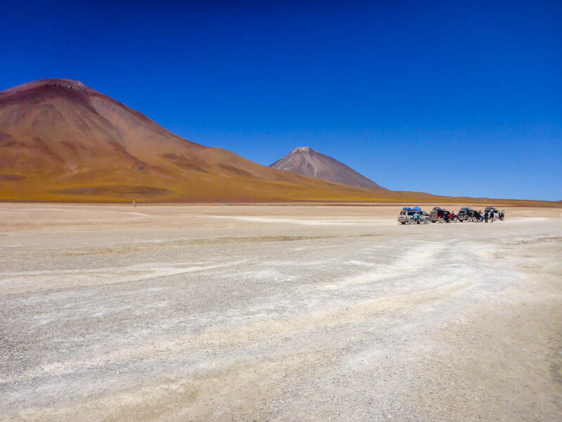 3 cars driving off in the distance on light beige colored sand with brown mountains in the background and bright blue sky