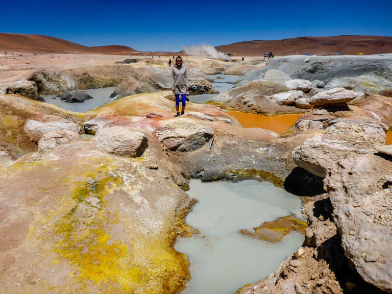 A woman standing on mutli-colored rock amidst several geysers in Sol de Mañana in Bolivia with steam vents, mud pools, hot springs, geysers and fumaroles.