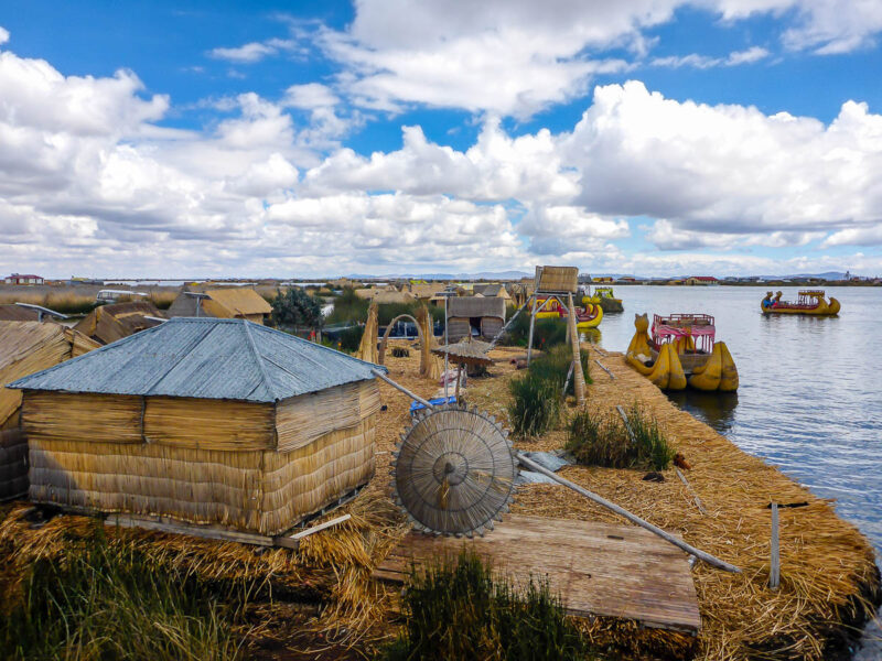 Uros floating islands made entirely of reeds on Lake Titicaca in Peru. A couple small shelters and boats with blue skies and clouds.