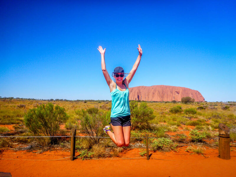 A woman jumping in the air with her hands above her head in excitement in front of the famous, Uluru or Ayers Rock in the Australian outback.