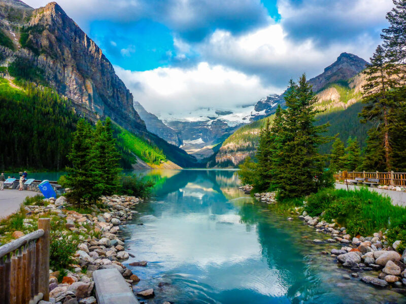 A mountain scene with turquoise lake in the middle, bright green pine trees on either side, snow capped mountains in the distance.