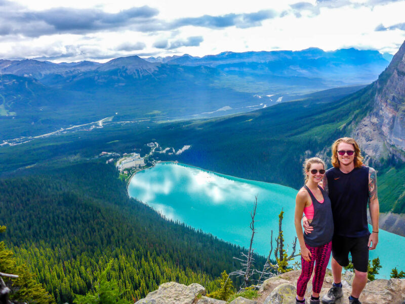 A woman and man standing at the top of the Beehive Hike overlooking Lake Louise with mountains and trees in the background