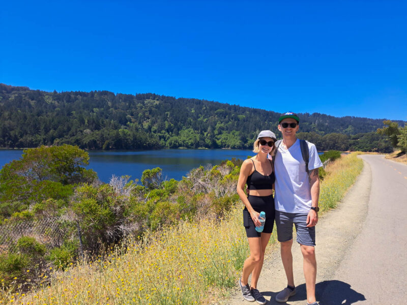 A man and woman posing at the side of a trail at a water reservoir with tall grass, plants, and trees around them. Rolling hills with green trees and bright blue sky in the background