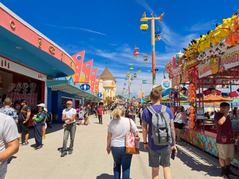 A man and woman walking away fro the camera along the Santa Cruz boardwalk with games, rides, and food stalls on either side of them