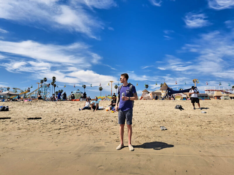 A man drinking a beer standing on sand in front of the Santa Cruz boardwalk with rides and games in the background.