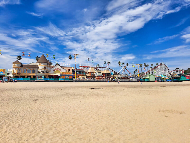 Santa Cruz boardwalk with games, rides, food kiosks and more set against a bright blue sky with whispy clouds and soft beige sand in front.