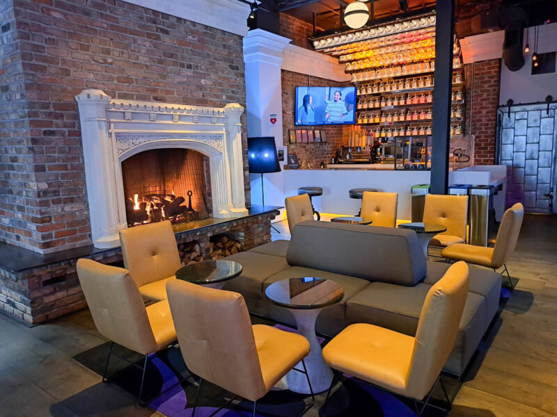 Lobby of hotel with a brick wall on one side, lit fireplace, dining table and leather chairs, comfortable seating, and a functioning bar with a tv and bottles of alcohol on the wall