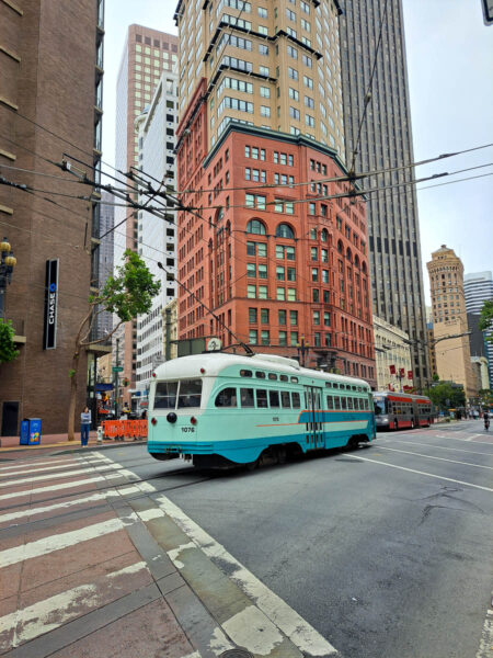 Turquoise colored cable car in the cross section of a busy street in San Francisco with cable lines above and tall buildings