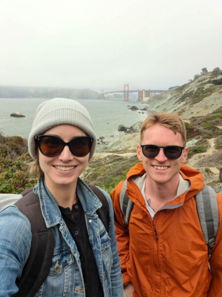 A man and woman smiling at the camera on a cloudy, foggy day in San Francisco with the Golden Gate Bridge shrouded in fog in the background along with the hillside and shubbery