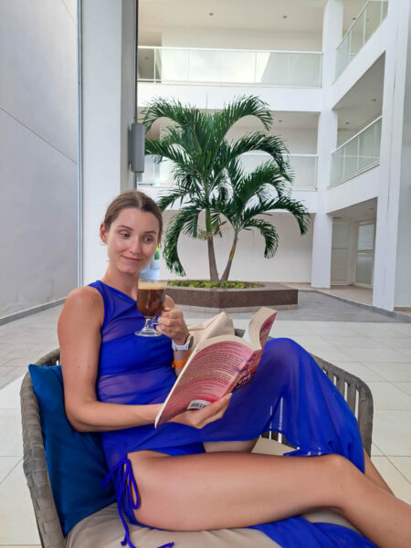 A woman dressed in a bright blue bikini and blue beach cover up sitting on a chair reading a book and drinking coffee, in an open air resort lobby with white walls behind and a big green tropical plant.