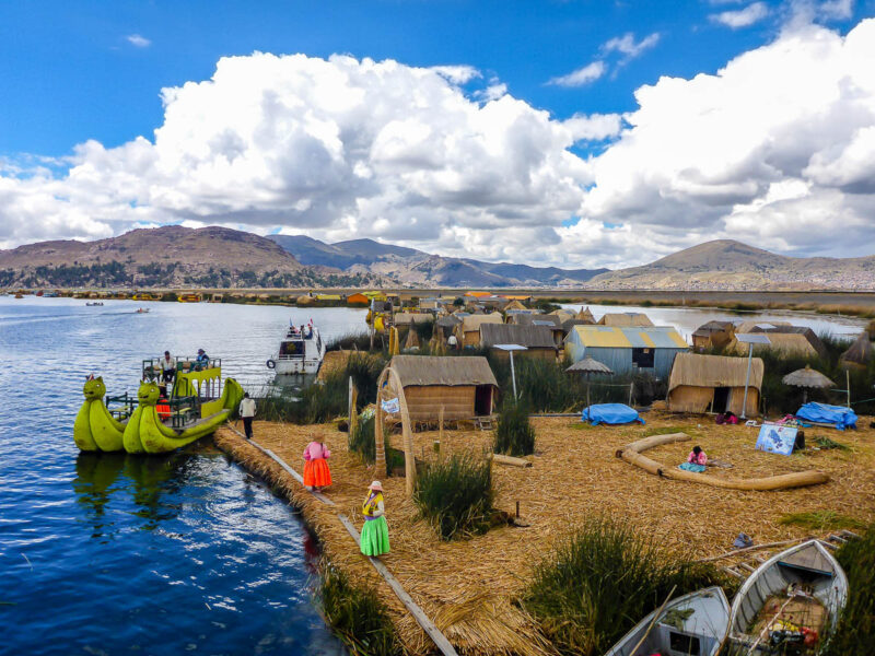 Floating reed islands with houses and structures all made of reeds, mountains, blue skies and white clouds in the background. Two women wearing colourful outfits standing near the edge or the island with a reed boat docked nearby.