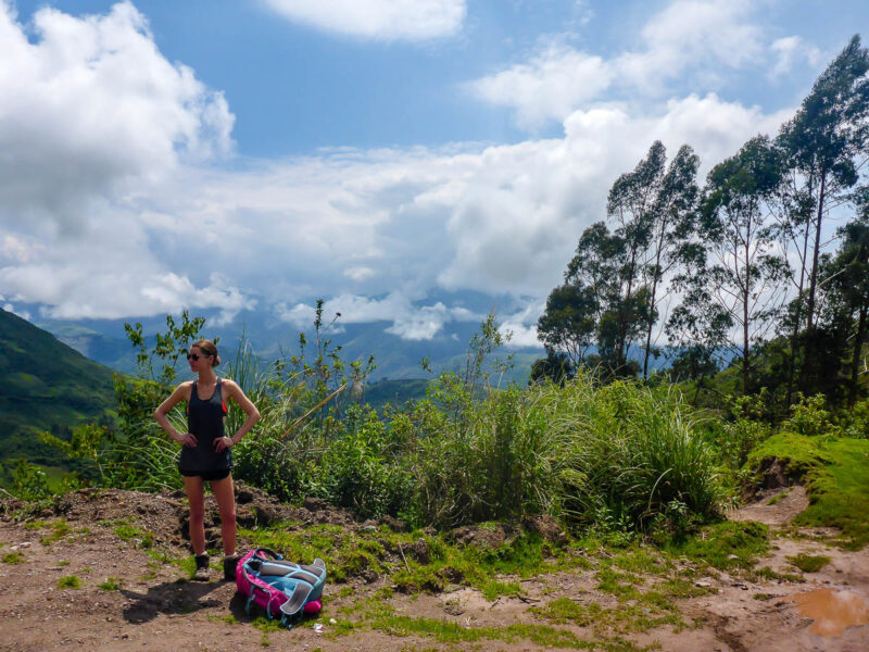 A woman standing on the side of a hiking trail with her backpack on the ground beside her, lush green plants and trees behind her, tall mountains in the background and blue skies with clouds.