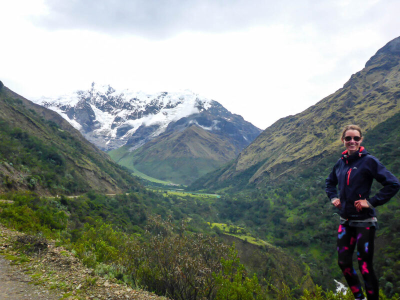 A woman posing in front of a view of Salkantay Trail with lush greenery and tall snow-capped mountains in the background