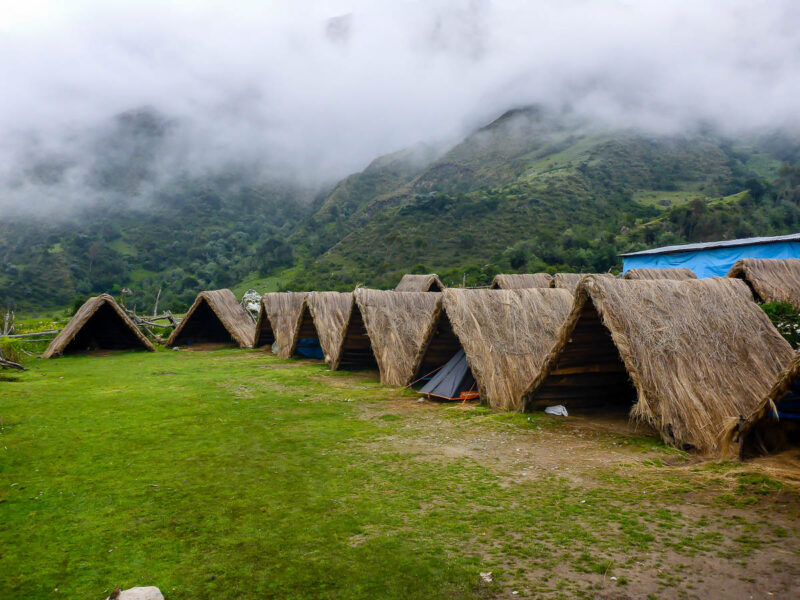 Several grass and wooden huts lined up in a row at Soraypampa campsite along the Salkantay Trail. Low-handing clouds and fog cover green-covered mountains behind.