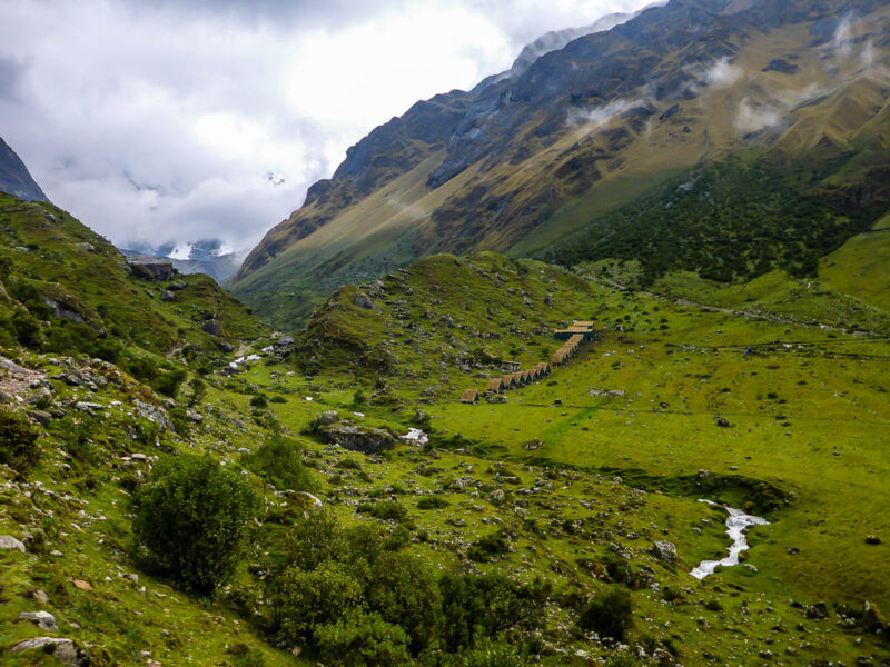 A view of green covered hills and mountains with a tiny village and camping huts in the valley. Tall mountains on either side and a cloudy sky.