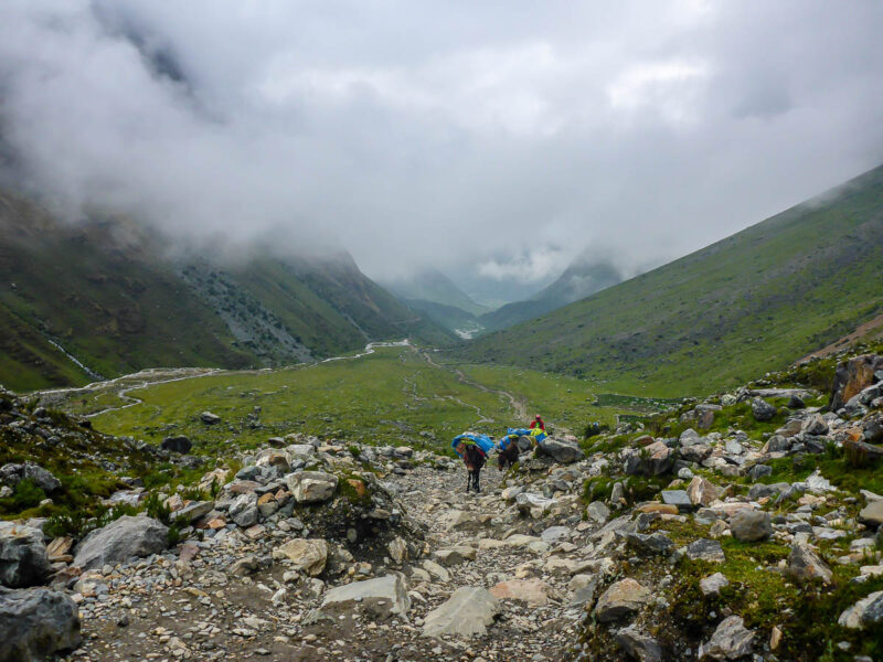 A few people trekking rocky portion of the Salkantay Trail with green grass and mountains.